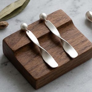 Op de afbeelding: Een paar zilverkleurige oorhangers met een parelstud en een gedraaid, langwerpig ontwerp. De oorbellen worden gepresenteerd op een houten blok, met een marmeren dienblad op de achtergrond. Een kleine zilveren zegel is ook zichtbaar.