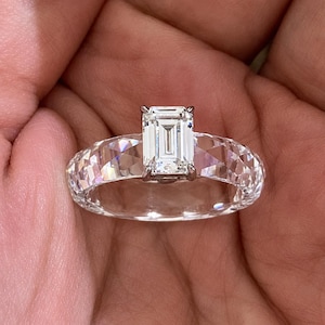 Könnte beinhalten: Ein klarer, facettierter Ring aus transparentem Material mit einem zentralen Smaragdschliff-Stein, der von einer silberfarbenen Metallfassung mit vier Krappen gehalten wird. Das Band hat eine geometrische, facettierte Oberfläche.