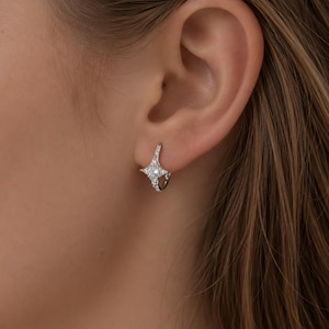 May include: A close-up of a silver hoop earring with a starburst design. The earring is encrusted with small, sparkling white stones. The earring is worn on an ear, showcasing the jewelry's design and sparkle.