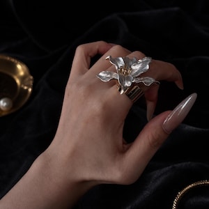 Può includere: Un anello a forma di fiore argentato con dettagli dorati. L'anello presenta un design floreale dettagliato con più petali e una fascia dorata. L'anello è indossato su un dito, mostrando le sue dimensioni e l'intricata lavorazione. Lo sfondo è un tessuto scuro e vellutato.