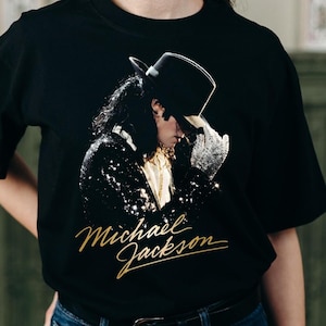 May include: Black t-shirt with a gold script "Michael Jackson" logo and a portrait of the singer wearing a top hat and sequined jacket. The image is a close-up of the singer's face and upper body.