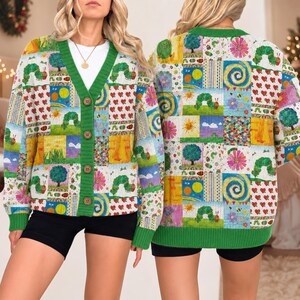 May include: A long-sleeve knit cardigan featuring a colorful patchwork pattern of The Very Hungry Caterpillar illustrations, including green caterpillars, suns, trees, and red hearts. The garment has a green V-neck collar and four brown buttons.