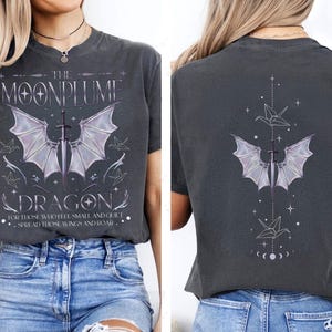 May include: Dark gray t-shirt with a graphic design featuring a sword, dragon wings, and the text "THE MOONPLUME DRAGON." The back of the shirt has a similar design. The shirt also has the quote: "FOR THOSE WHO FEEL SMALL AND QUIET... SPREAD THOSE WINGS AND ROAR."