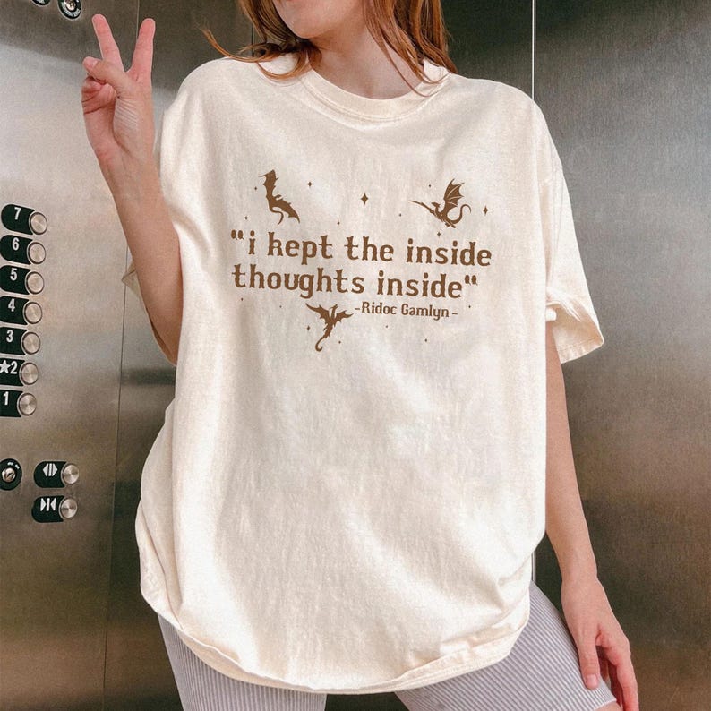 May include: Cream-colored t-shirt with the quote "i kept the inside thoughts inside" in brown text. The shirt features dragon illustrations. The quote is attributed to Ridoc Gamlyn.