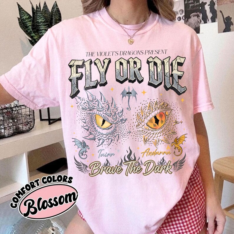 May include: A light pink t-shirt featuring a graphic print with the text THE VIOLET'S DRAGONS PRESENT FLY OR DIE and Brave The Dark. The design includes two detailed dragon eyes, small dragon illustrations, and flame motifs in shades of yellow, grey, and black.