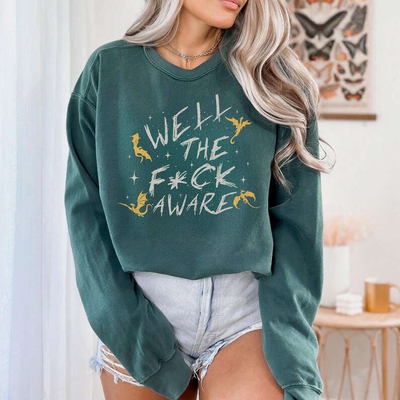 May include: Dark green sweatshirt with the text "WELL THE F*CK AWARE" in a whimsical font. The design includes small star and dragon illustrations in gold. The sweatshirt is a casual, comfortable style.