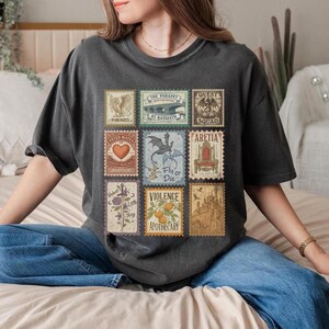 May include: Charcoal grey t-shirt with a collection of vintage postage stamp designs. The stamps showcase illustrations such as a heart, a dragon, and a castle. Text on the stamps includes "THE PARAPET", "QUEST", "Fly or Die", and "VIOLENCE".