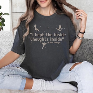 May include: Dark gray t-shirt with the quote "I kept the inside thoughts inside" and dragon illustrations. The quote is attributed to Ridoc Gamlyn. The shirt is a crew neck and has short sleeves.