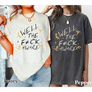 May include: Ivory and pepper-colored t-shirts with the text "WELL THE F*CK AWARE" in black, with gold dragon and star graphics. The shirts are short-sleeved and have a relaxed fit.