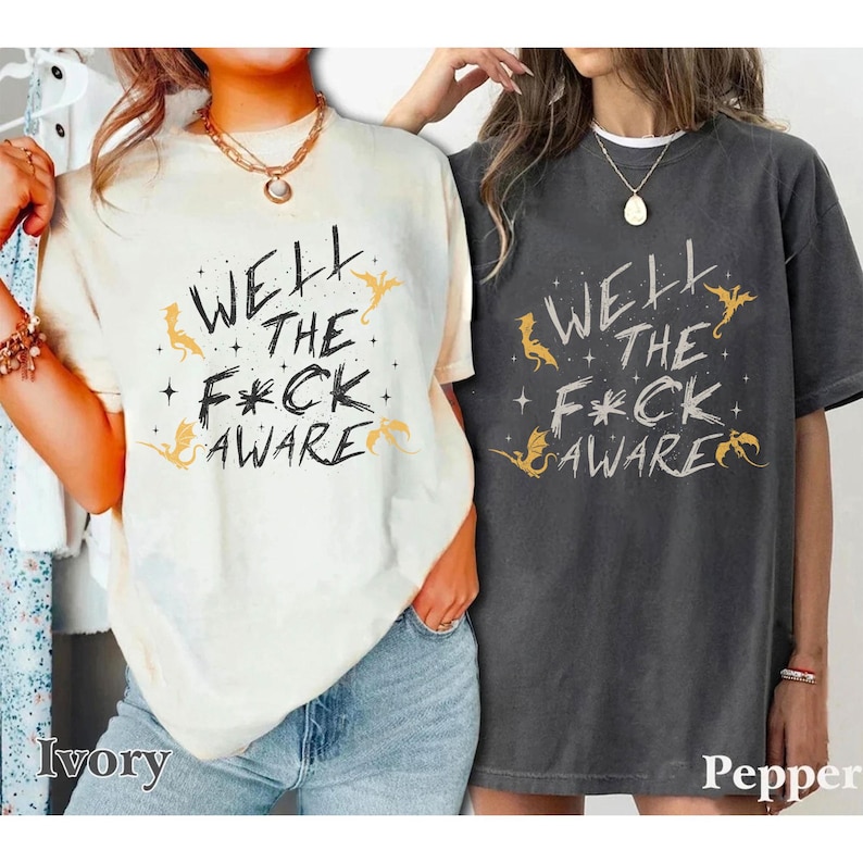 May include: Ivory and pepper-colored t-shirts with the text "WELL THE F*CK AWARE" in black, with gold dragon and star graphics. The shirts are short-sleeved and have a relaxed fit.
