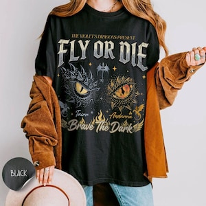 May include: A black t-shirt featuring the text THE VIOLET'S DRAGONS PRESENT FLY OR DIE and BRAVE THE DARK. The graphic includes two detailed dragon eyes, one blue and one orange, surrounded by small dragon silhouettes, stars, and flames.