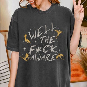 May include: Dark gray t-shirt with the text "Well the F*ck Aware" in a distressed font, accompanied by gold dragon illustrations and stars. The shirt has a relaxed fit.