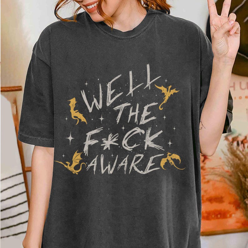 May include: Dark gray t-shirt with the text "Well the F*ck Aware" in a distressed font, accompanied by gold dragon illustrations and stars. The shirt has a relaxed fit.