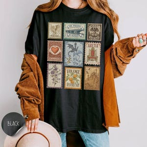 May include: Black t-shirt with nine vintage-style postage stamp designs. Each stamp features a unique illustration and text, such as a heart, dragon, and castle. The shirt has a classic crew neck and short sleeves.