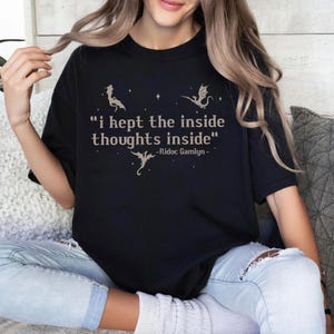 May include: Black t-shirt with the quote "i kept the inside thoughts inside" and dragon illustrations. The quote is in a serif font, with the author's name, Ridoc Gamlyn, below. The shirt is a comfortable, casual style.