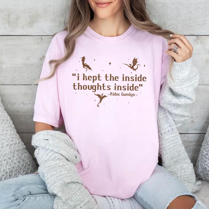 May include: A light pink t-shirt with the quote "I kept the inside thoughts inside" in brown text, along with dragon illustrations. The quote is attributed to Ridoc Gamlyn. The shirt is a casual wear item.