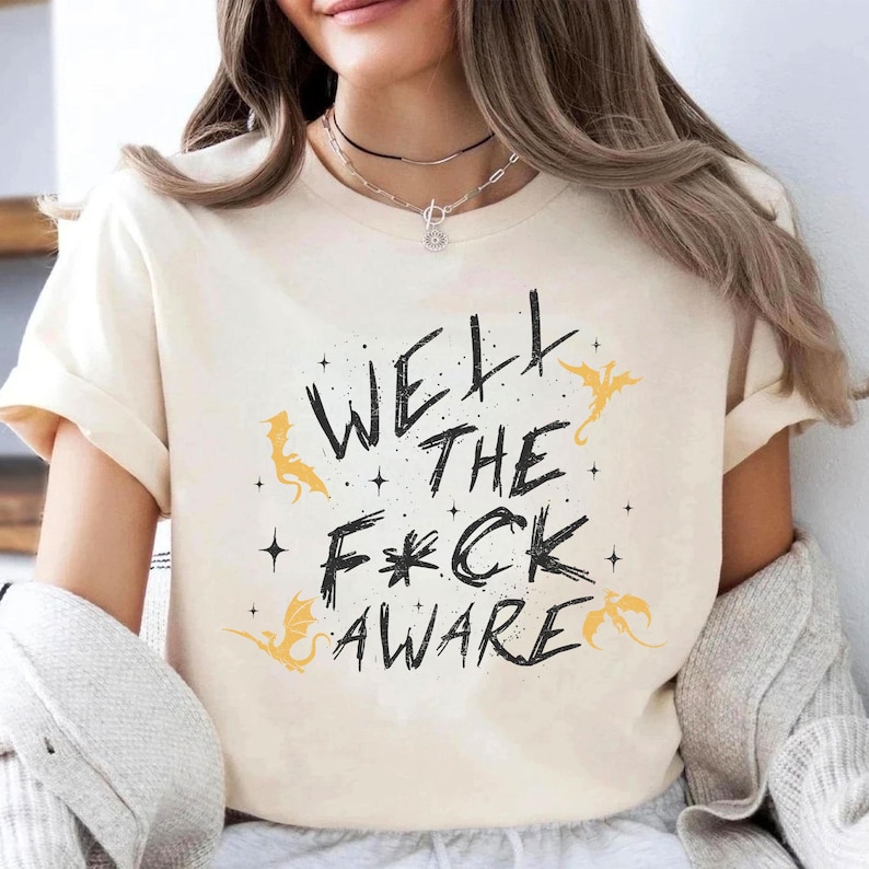 May include: Cream-colored t-shirt with the text "WELL THE F*CK AWARE" in black, with yellow dragon illustrations. The shirt has short sleeves and a casual fit. The design includes stars and dragon silhouettes.