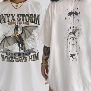 May include: Cream-colored t-shirt featuring a black dragon graphic with the text "ONYX STORM" and "BRAVE THE DARK". Below the dragon is the text "WILL NOT DIE TO DAY I WILL SAVE HIM". The back of the shirt has a matching dragon and celestial design.
