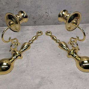 May include: A pair of polished brass wall sconces featuring a curved arm, a circular candle cup, and a decorative elongated base with a mounting hole at the top. The metal surface is reflective and smooth with a bright gold-toned finish.