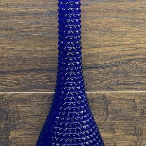 May include: A tall, cobalt blue glass vase with a textured, diamond-like pattern. The vase has a narrow neck and a wider, teardrop-shaped base. The vase is approximately 12 inches tall.