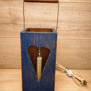May include: A rectangular blue wooden lantern with a heart-shaped cutout on the front, featuring a wire mesh backing and a central electric candle bulb. The lantern includes a brown wooden handle at the top and a white power cord with a switch.