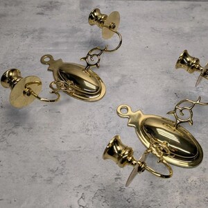 May include: A pair of polished brass wall sconces, each featuring an oval backplate with a hanging loop at the top. Two curved arms extend from each base, supporting candle cups with circular drip pans. The metal has a reflective gold finish.