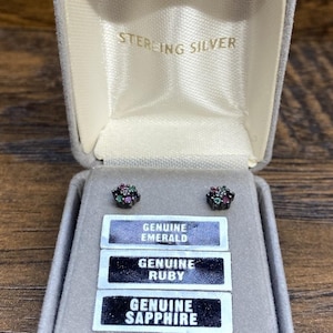 May include: A gray jewelry box with the words "STERLING SILVER" printed on the inside lid. The box contains two small stud earrings with a cluster of gemstones. Below the earrings are three labels that read "GENUINE EMERALD", "GENUINE RUBY", and "GENUINE SAPPHIRE".