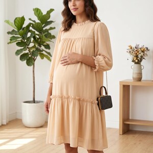 May include: A knee-length, light beige dress with long sleeves, a ruffled neckline, and a ruffled hem. The dress features a loose, flowing silhouette with gathered fabric at the chest and a small, dark brown, box-shaped handbag with a gold chain strap.