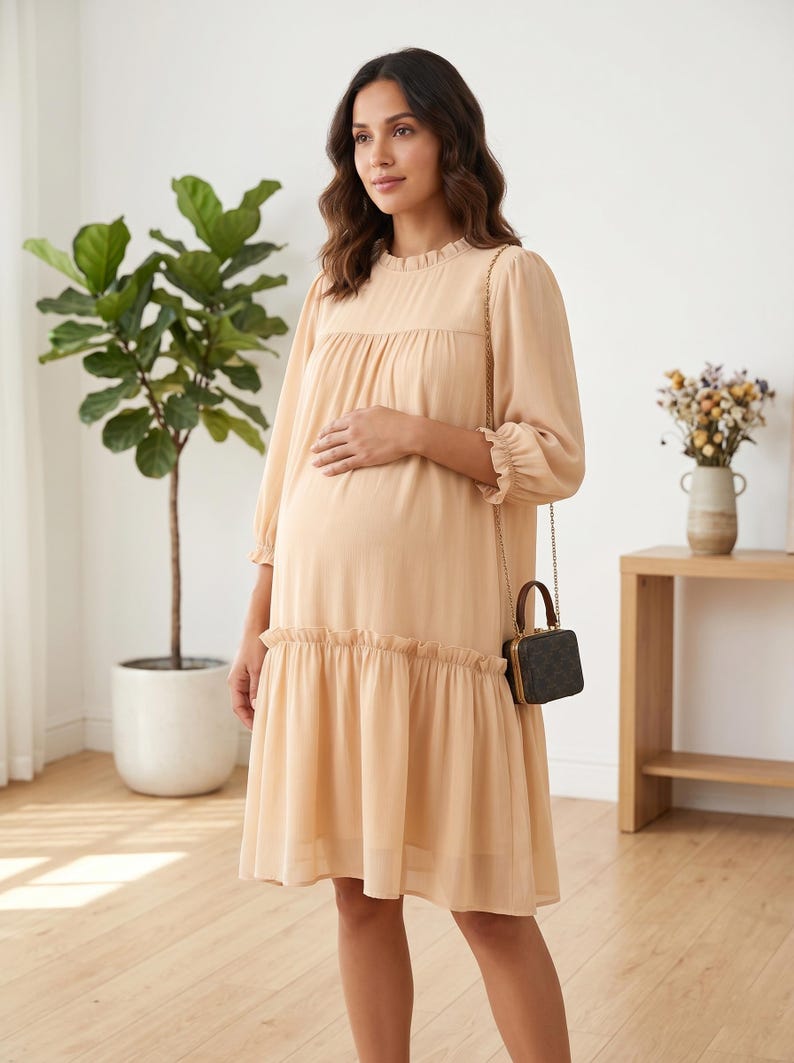 May include: A knee-length, light beige dress with long sleeves, a ruffled neckline, and a ruffled hem. The dress features a loose, flowing silhouette with gathered fabric at the chest and a small, dark brown, box-shaped handbag with a gold chain strap.