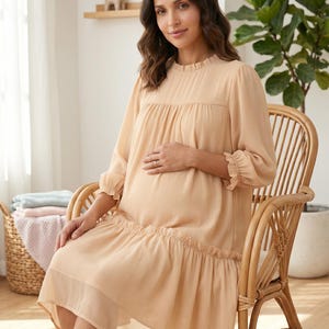 May include: A knee-length, light beige dress with long sleeves, a ruffled mock neckline, and a gathered drop-waist skirt. The dress features sheer, lightweight fabric with a subtle texture, ruffled sleeve cuffs, and a loose, flowing silhouette.