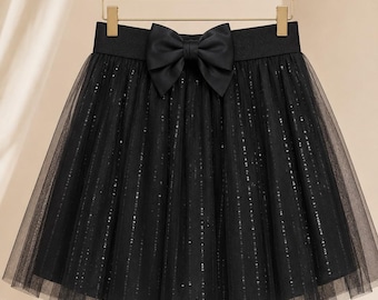 May include: A black tulle skirt featuring a wide elastic waistband adorned with a large, centered black satin bow. The skirt is constructed with multiple layers of sheer black tulle fabric, embellished with vertical rows of shimmering silver sequins.