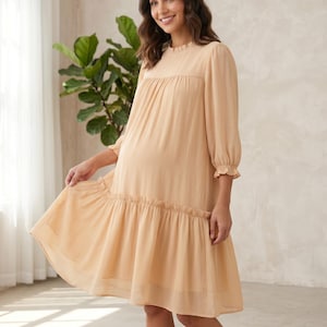 May include: A knee-length, light beige dress featuring a high, ruffled neckline, three-quarter length sleeves with ruffled cuffs, and a gathered drop-waist skirt. The garment is made of a semi-sheer, lightweight fabric with a subtle vertical texture.