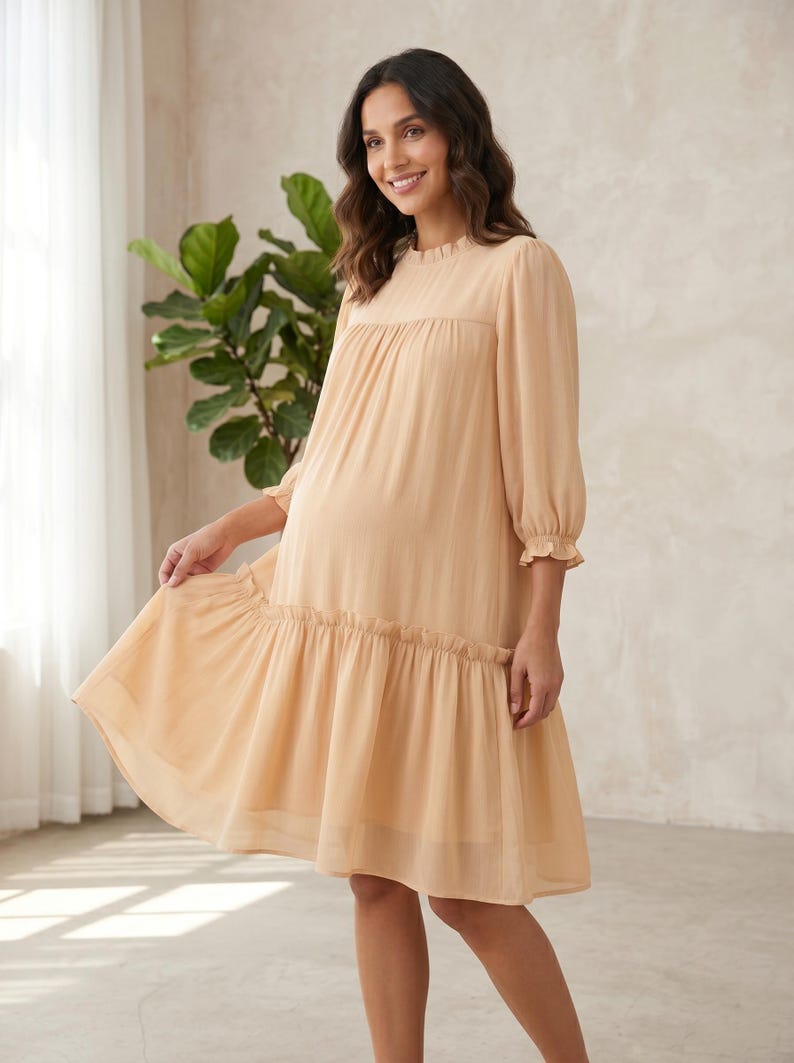 May include: A knee-length, light beige dress featuring a high, ruffled neckline, three-quarter length sleeves with ruffled cuffs, and a gathered drop-waist skirt. The garment is made of a semi-sheer, lightweight fabric with a subtle vertical texture.
