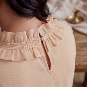 May include: A light peach-colored blouse featuring a high ruffled collar with a smocked texture. The back of the garment includes a keyhole opening secured by a single round white faux pearl button with a metallic gold-toned rim detail.