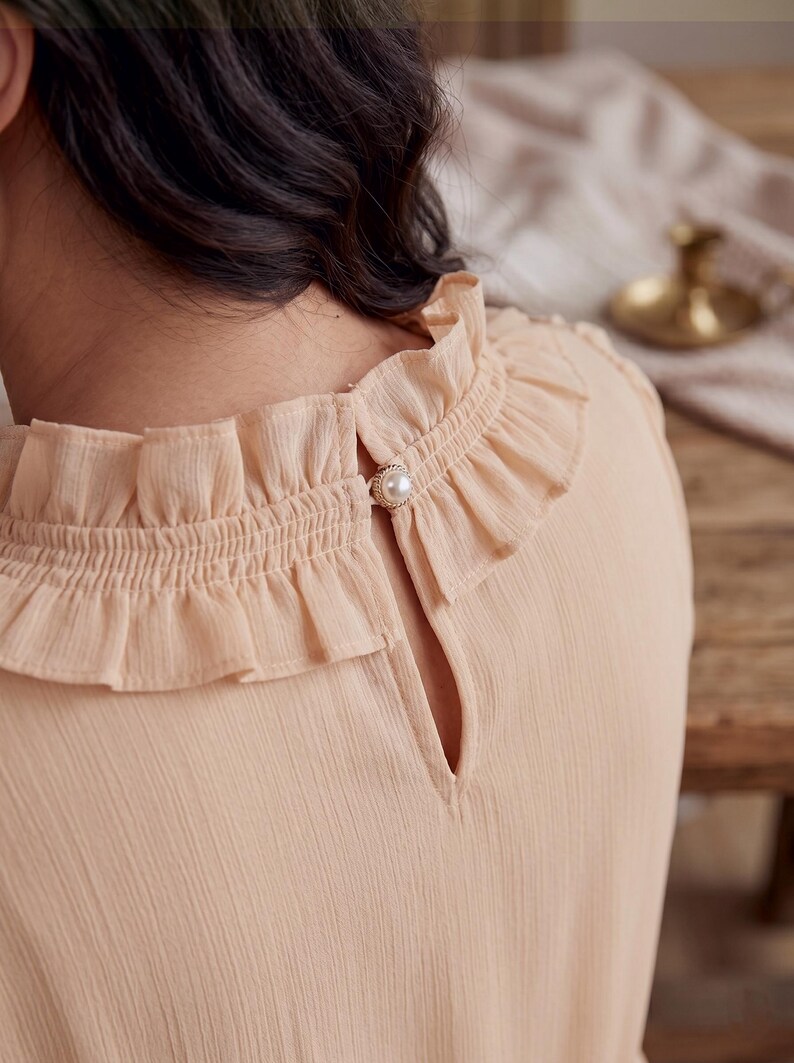 May include: A light peach-colored blouse featuring a high ruffled collar with a smocked texture. The back of the garment includes a keyhole opening secured by a single round white faux pearl button with a metallic gold-toned rim detail.