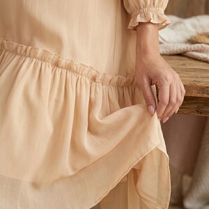 May include: A light beige dress featuring long sleeves with gathered, ruffled cuffs and a tiered skirt design. The fabric has a subtle vertical texture, and the hemline is accented with a wide, gathered ruffle detail that adds volume to the garment.