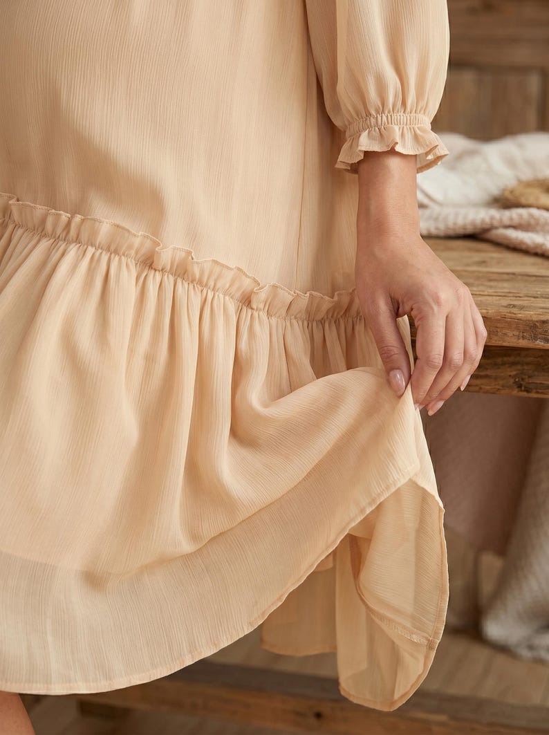May include: A light beige dress featuring long sleeves with gathered, ruffled cuffs and a tiered skirt design. The fabric has a subtle vertical texture, and the hemline is accented with a wide, gathered ruffle detail that adds volume to the garment.