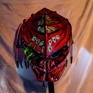 May include: A red and black patterned helmet featuring a series of glowing green symbols across the top and side. The helmet has a pointed, angular design with multiple black, spike-like appendages extending from the sides and back of the structure.