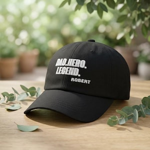 Könnte beinhalten: Schwarze Baseballkappe mit dem weißen Text "DAD. HERO. LEGEND. ROBERT". Die Kappe hat einen gebogenen Schirm und ein klassisches Sechs-Panel-Design. Der Hut ist aus strapazierfähigem Stoff gefertigt und für den täglichen Gebrauch geeignet.