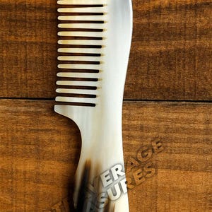 May include: A handcrafted comb with a curved handle and fine teeth. The comb is made of a light-colored material with dark brown streaks near the base. The comb is approximately 7 inches long.