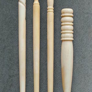 May include: Four light-colored wooden crochet hooks arranged vertically. The leftmost hook features a spiral carved handle, while the other three hooks have varying ribbed or grooved handle designs. Each tool ends in a distinct hook shape.