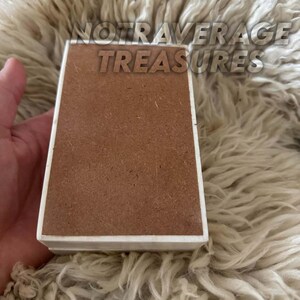 May include: A rectangular box with a smooth white border and a flat, textured brown center panel. The text NOTAVERAGE TREASURES is printed in a bold, sans-serif font across the top portion of the object, which has a clean, geometric design.
