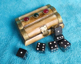 May include: A small, arched-top wooden chest with a light tan finish, featuring ornate gold-toned metal trim, a central latch, and three embedded round cabochon stones in red and green. Five black six-sided dice with white pips are arranged nearby.