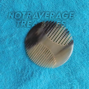 Puede incluir: Un peine para el cabello redondo, liso y semitranslúcido que presenta un degradado de colores desde el marrón oscuro hasta el crema con dientes finos y espaciados uniformemente. El texto NOTRAVERAGE TREASURES aparece sobre el peine.