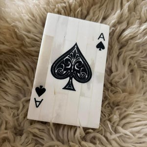 May include: A rectangular decorative object crafted from white inlaid segments, designed to resemble an ace of spades playing card. The surface features a large central black spade symbol with intricate filigree details, accompanied by a smaller spade icon and the letter A in the top right and bottom left corners.
