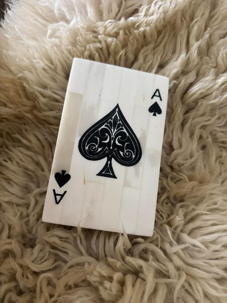 May include: A rectangular decorative object crafted from white inlaid segments, designed to resemble an ace of spades playing card. The surface features a large central black spade symbol with intricate filigree details, accompanied by a smaller spade icon and the letter A in the top right and bottom left corners.