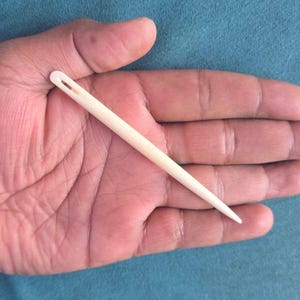 May include: A light-colored, wooden needle with a large eye. The needle is held in a hand against a blue background. The needle is tapered to a sharp point.