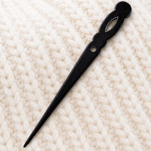 Pode incluir: Um palito de cabelo de madeira preta com um topo decorativo e uma extremidade pontiaguda. O acessório de cabelo é mostrado contra um fundo texturizado creme.