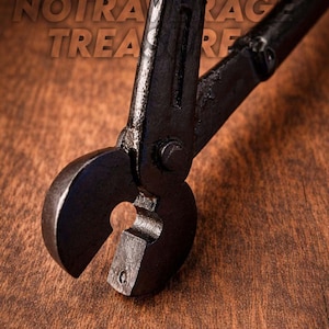 Può includere: Primo piano di uno strumento in metallo nero con una testa curva e un'apertura a forma di buco della serratura. Lo strumento è posto su uno sfondo di legno. Il testo "NOTRA TREASURE" è visibile sullo sfondo.