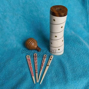 May include: A cylindrical white container with a dark wood top, featuring a spiral pattern of small gold dots. Beside it sits a small, round, brown wooden spinning top and four thin, flat, cream-coloured sticks with red and brown geometric designs.